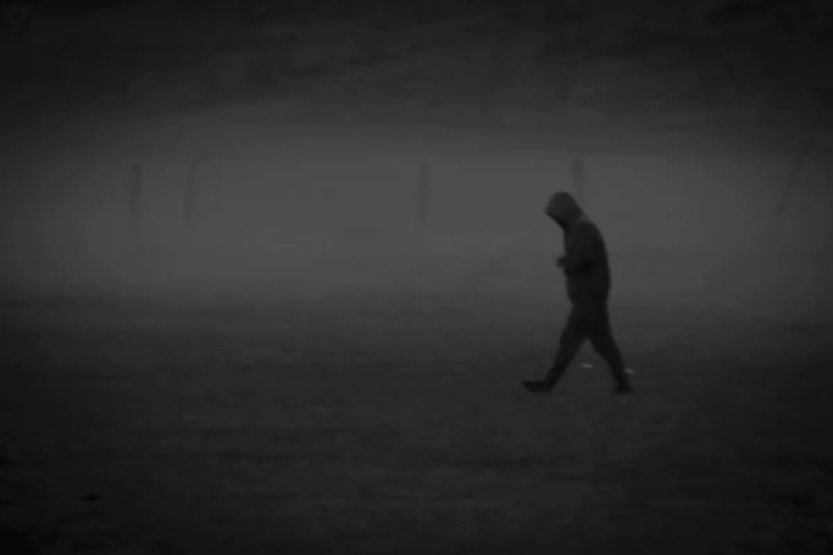 Hooded figure walking through dense fog across an empty field